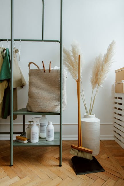 A minimalist metal clothing rack positioned against a plain white wall in a well-lit room. The rack holds a few hangers with clothing, including a beige coat and a green garment. On the middle shelf, 