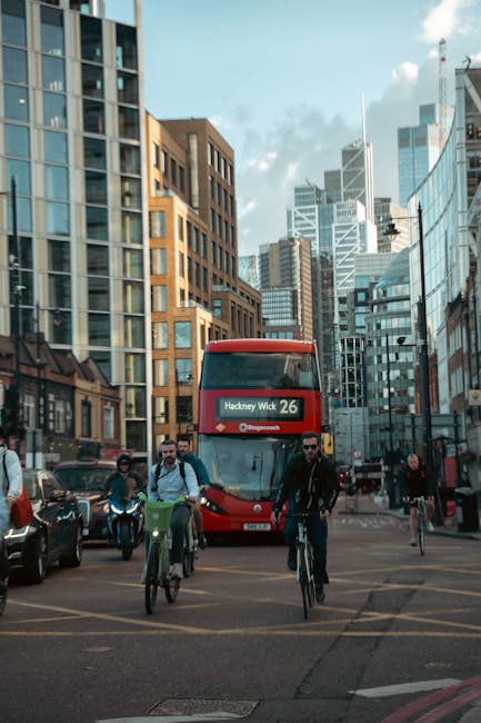 A busy urban street scene in Hackney with high-rise buildings and modern glass-fronted offices. In the foreground, several pedestrians, some on bikes and others walking, are crossing the street. A red