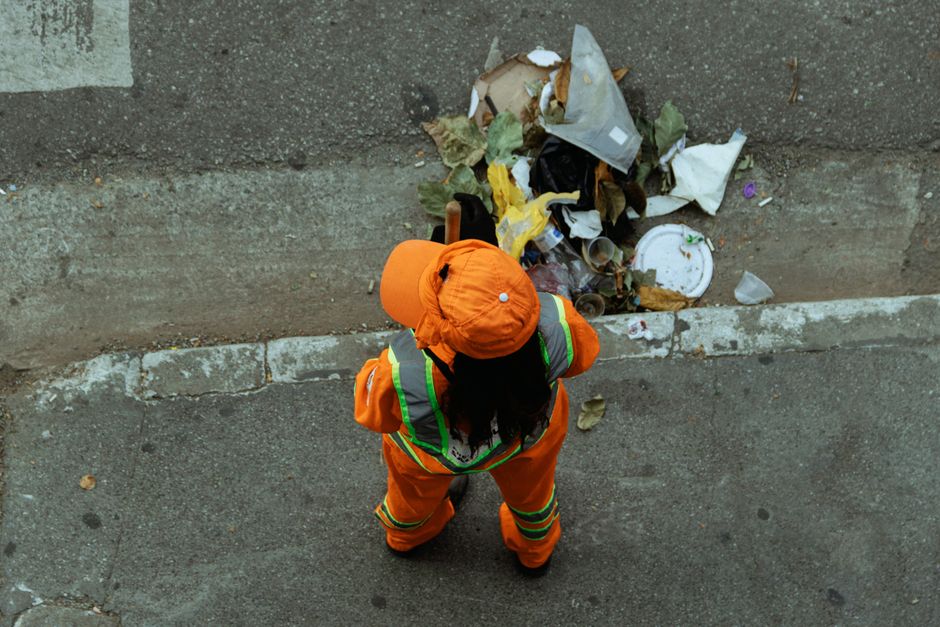 A person wearing a high-visibility orange safety vest, black gloves, and a clear plastic raincoat is pushing a green cleaning cart equipped with three compartments, containing a broom, mop, and cleani