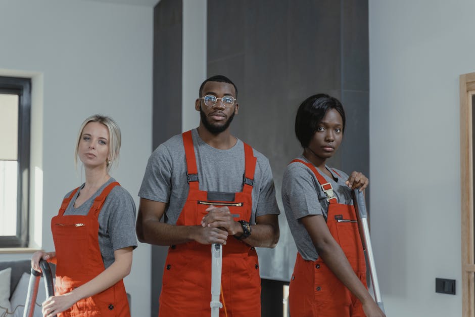 Two professional cleaners from Cleaners Hackney are performing surface cleaning in a modern living room. One cleaner, on the left, is vacuuming the wooden floor with a portable upright vacuum cleaner,