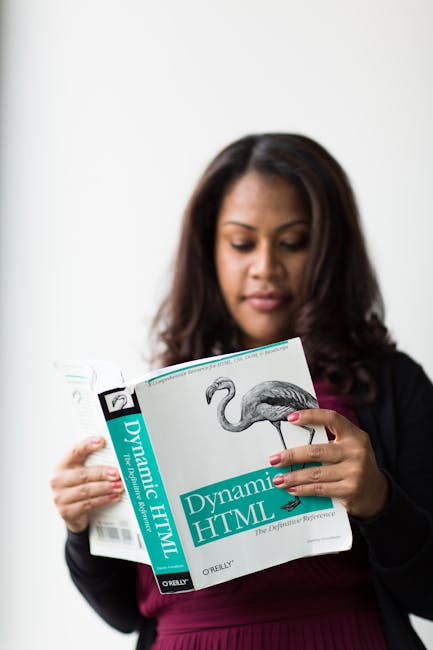 A woman with long dark hair, dressed in a black blazer and maroon top, is holding an open technical book titled 'Dynamic HTML' with a cover illustration of a flamingo. She appears to be reading or stu